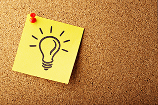 Yellow sticky note with a lightbulb idea drawing pinned to a cork board.
