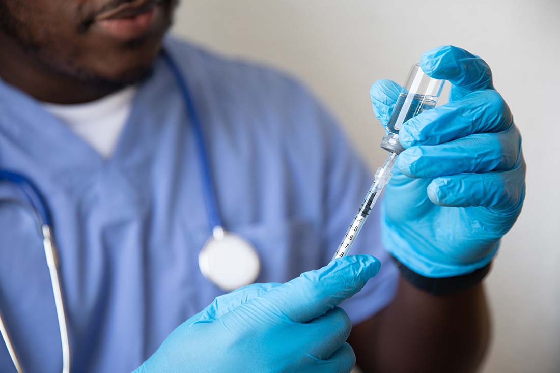 Male nurse with blue scrubs, stethoscope around his neck, and rubber gloves drawing a vaccine.