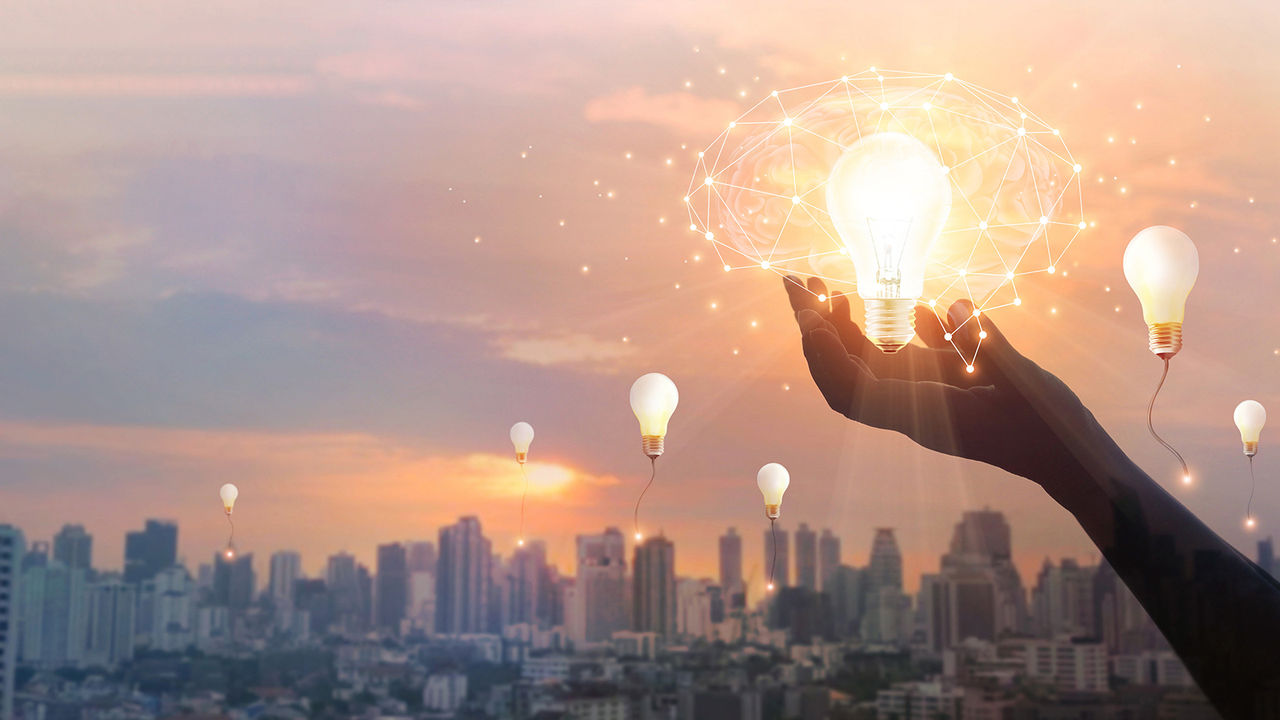 Hand holding lightbulb emitting neural network late with more lightbulbs and city skyline background.