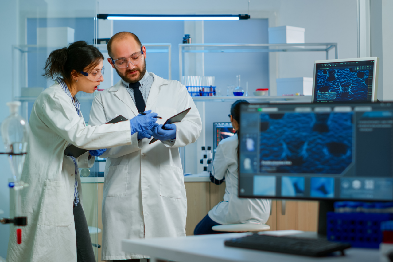 Modern Medical Research Laboratory: Two Scientists Working, Using Digital Tablet, Analyzing Test, Talking. Advanced Scientific Pharmaceutical Lab for Medicine, Biotechnology Development.